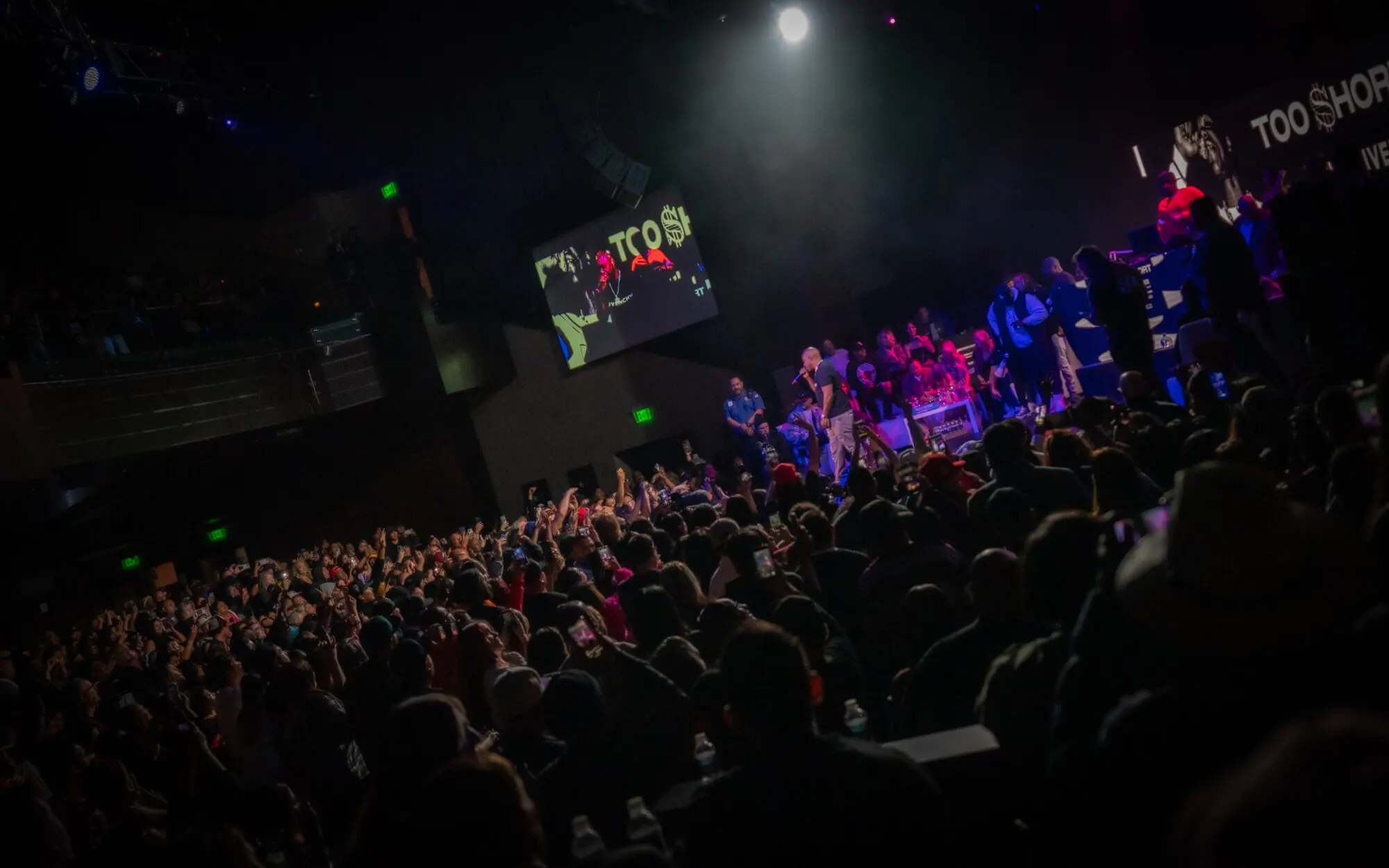 The room from the floor, phones up, the stage lit at the far end under the screens
