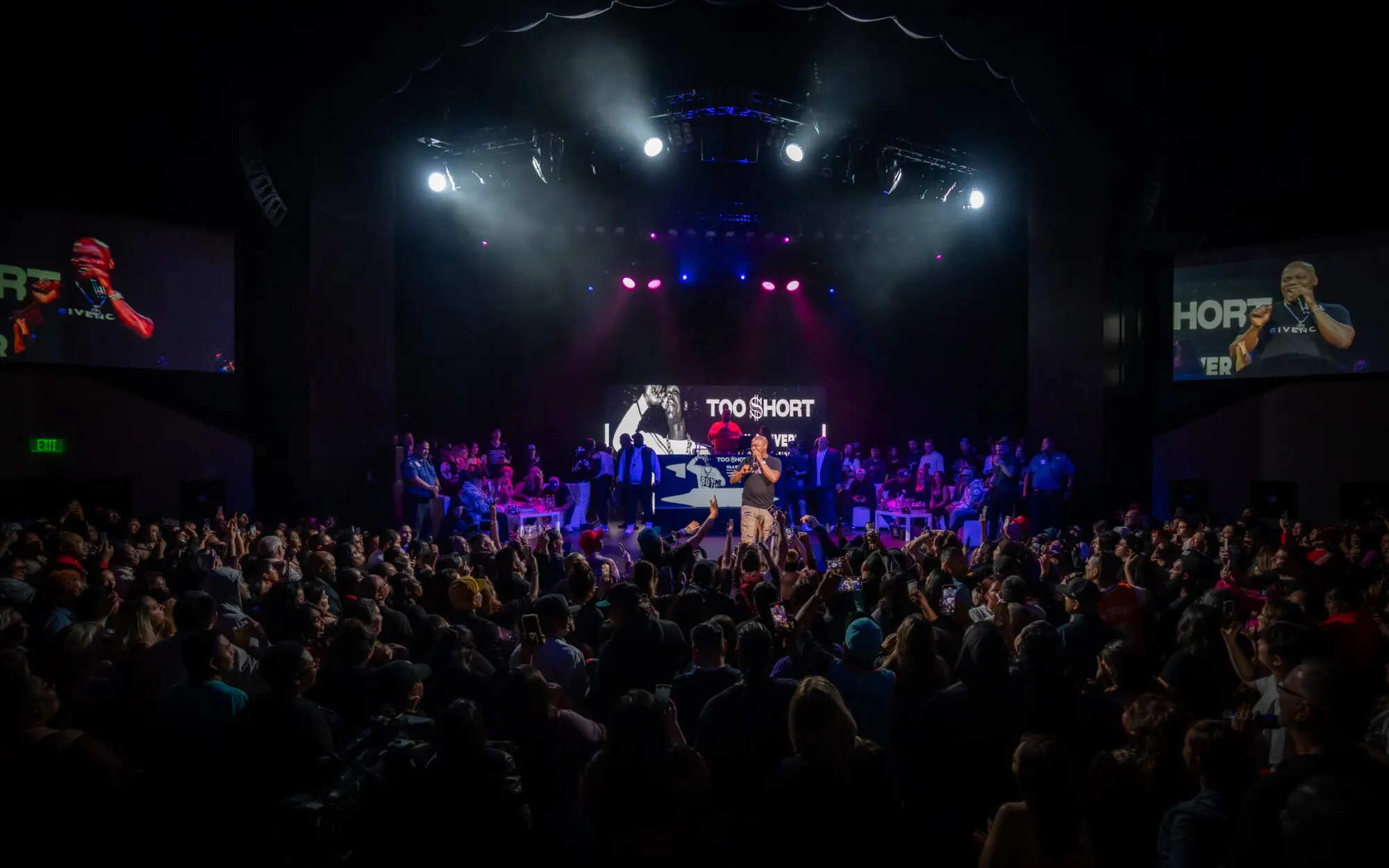 The room from the back, the crowd facing a lit stage with the screens either side
