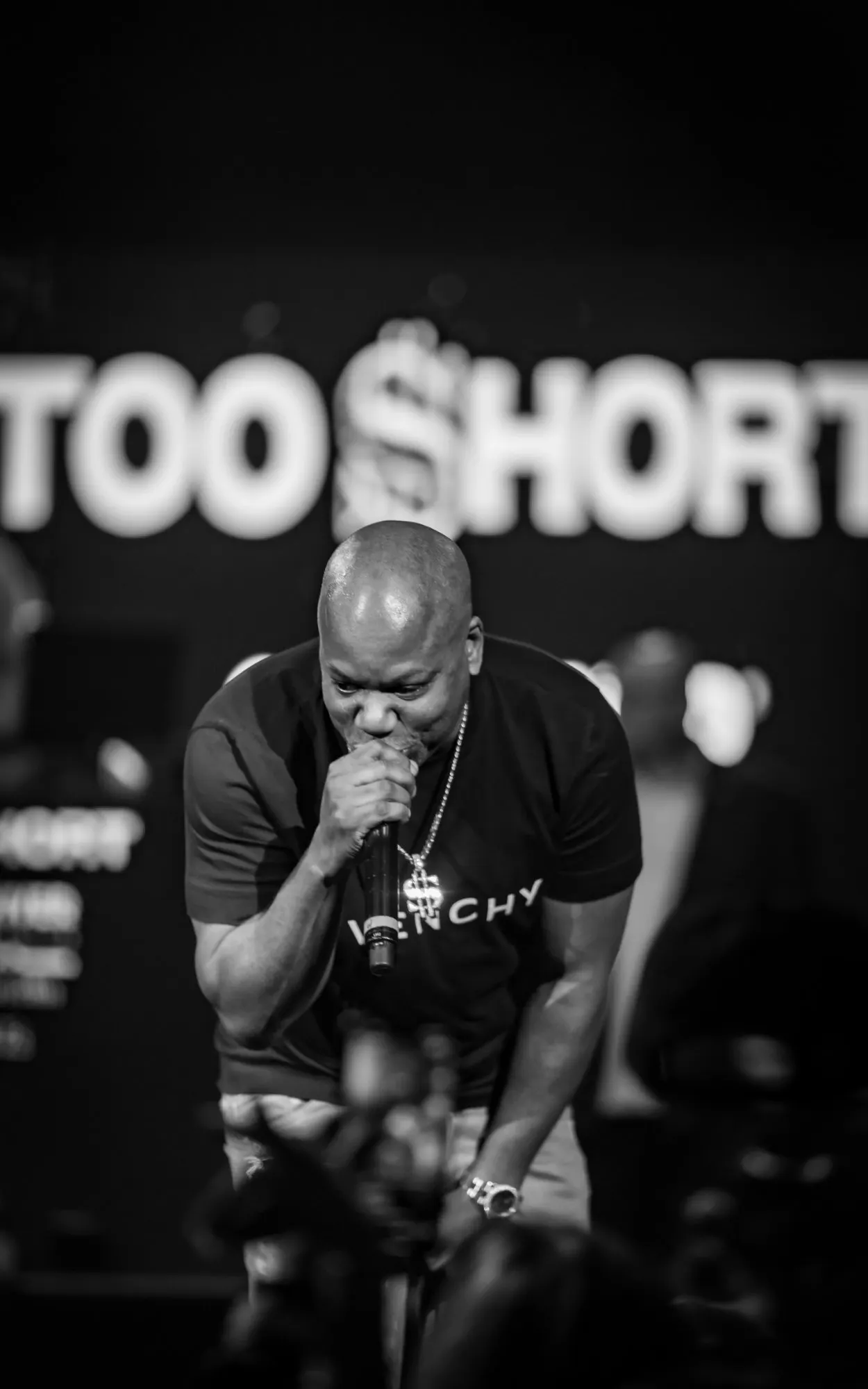 A performer bent low over the microphone in black and white, the lit stage banner behind him
