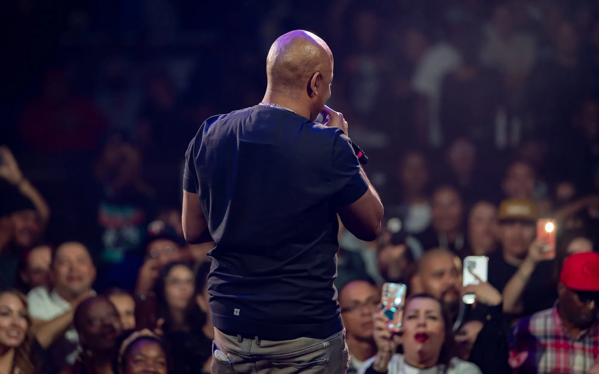 A performer facing the crowd from the stage, the front rows lit and phones raised