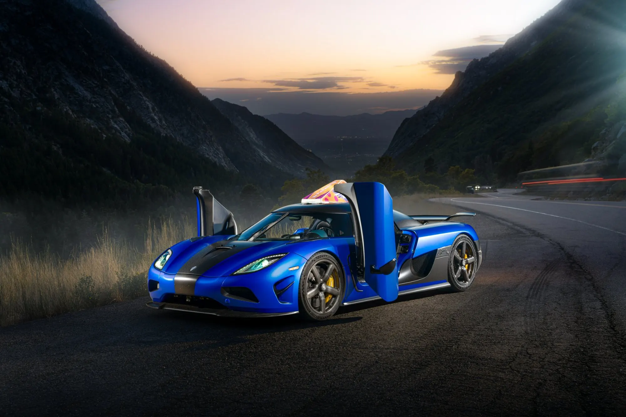 Blue hypercar on the mountain road with its doors raised, the valley behind it